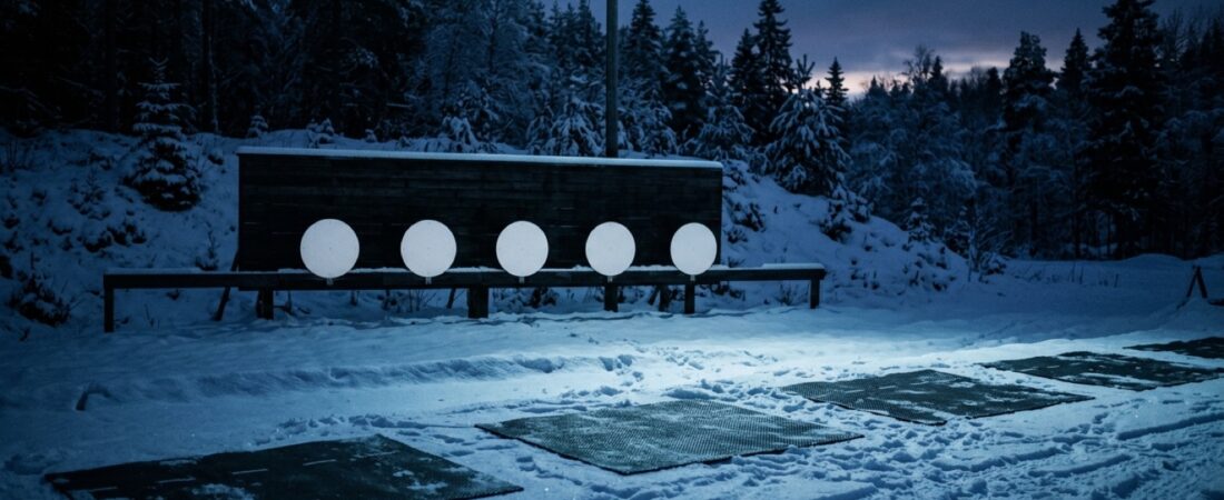 Hämärässä valaistu ampumahiihdon ampumarata lumisateessa, viisi taulua rivissä ja tyhjät ampumamatot etualalla.