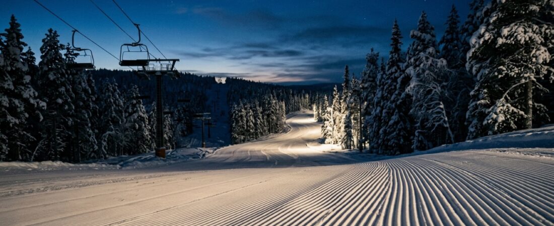 Tyhjä muokattu laskettelurinne yöllä, rinnevalot valaisevat lunta ja taustalla lumiset kuuset sinertävässä hämärässä.