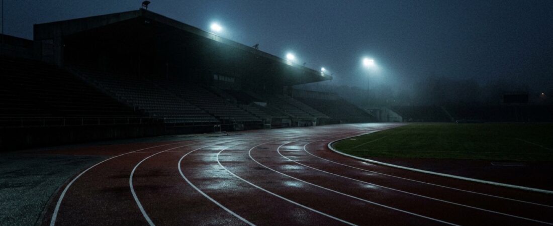 Tyhjä yleisurheilustadion hämärässä, sinisen juoksuradan kaarre ja valonheittimien sähkönsininen hehku.